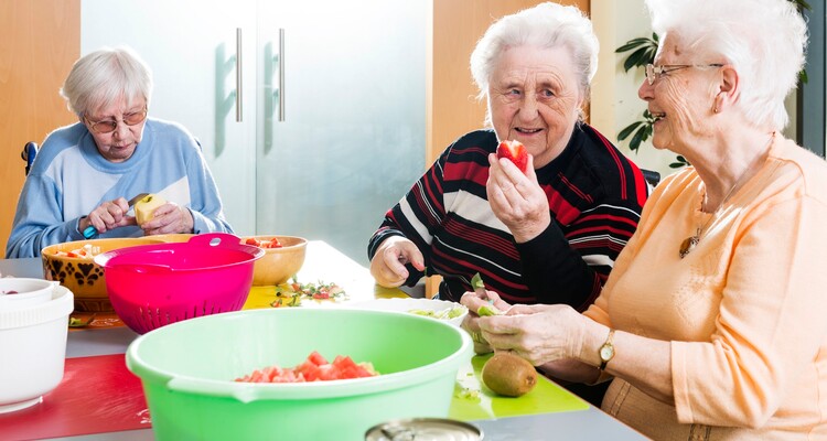 Drei fröhliche Bewohnerinnen sitzen nebeneinander und schneiden Obst.